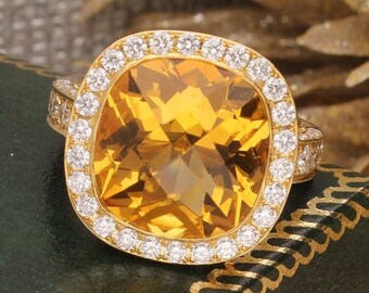 Pode incluir: Um anel de ouro com uma grande gema amarela de corte almofada, cercada por um halo de gemas menores, redondas e brancas. O anel é apresentado numa caixa de veludo verde.
