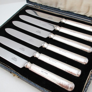 May include: A set of six silver knives in a blue box with a black velvet interior. The knives have shiny silver blades and textured handles. The box is open, revealing the knives neatly arranged.