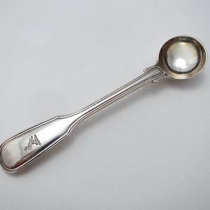 Peut inclure: Une cuillère en argent avec un bol rond et un long manche fin. La poignée a un design décoratif et un petit emblème près de l'extrémité. La cuillère est probablement destinée à servir ou à manger, et elle est en métal brillant et réfléchissant.