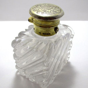 May include: A clear, square-shaped perfume bottle with a decorative, silver-colored lid. The bottle has a textured, cut-glass design. The lid is ornate with a floral pattern and a gold-colored band. A vintage-style item.