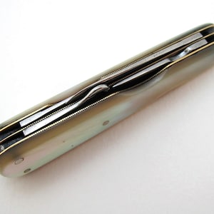 May include: A closed pocket knife with a mother-of-pearl handle. The knife has multiple blades and a gold-coloured trim. The handle is pearlescent with a slight iridescence. The knife is lying on a white surface.