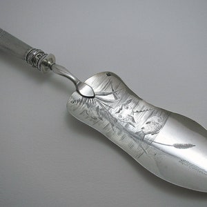 Op de afbeelding: Een zilveren taartschep met een sierlijk handvat en een blad gegraveerd met een scène van vissen en riet.