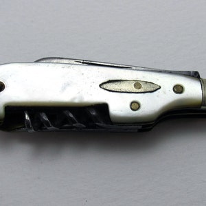 Puede incluir: Un cuchillo de bolsillo cerrado con un mango de nácar blanco y múltiples hojas. El cuchillo tiene un acento de latón y una pequeña letra 'G' en el extremo.