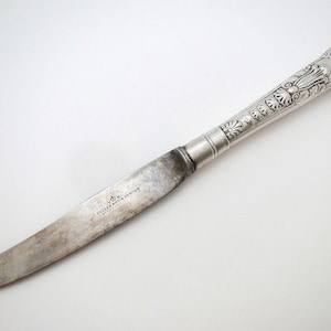 Puede incluir: Un cuchillo de mantequilla plateado con un mango ornamentado. El mango presenta diseños florales y de volutas detallados. La hoja muestra signos de desgaste y tiene texto cerca de la base. Una pieza clásica de cubertería.