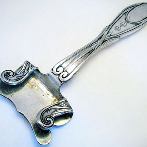 Może przedstawiać: Srebrny nóż do sera w stylu Art Nouveau. Rękojeść ma owalny kształt z kwiatowymi detalami. Ostrze ma charakterystyczny zakrzywiony kształt z dekoracyjnymi zawijasami. Nóż jest przeznaczony do serwowania sera.