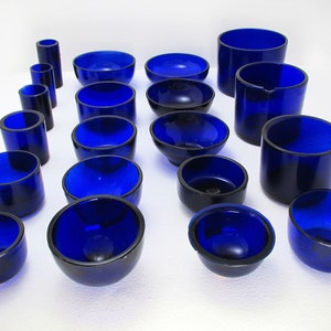 Puede incluir: Un conjunto de 17 cuencos y cilindros de vidrio azul cobalto de diferentes tamaños. Los cuencos son redondos y tienen un acabado liso y pulido. Los cilindros son altos y tienen un labio ligeramente acampanado.