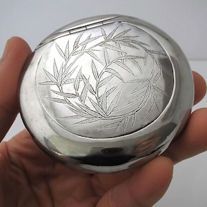 Puede incluir: Un estuche compacto redondo de plata con tapa abisagrada. La parte superior está grabada con un diseño detallado de hojas de bambú. El estuche compacto se sostiene en una mano, mostrando su superficie pulida y reflectante. Un estilo clásico y elegante.