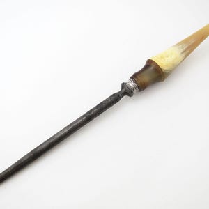 Könnte beinhalten: Eine lange, dunkle Haarspange aus Metall mit einem dekorativen Griff. Der Griff hat eine hellbraune und cremefarbene Hornspitze, einen dunkelbraunen Abschnitt und ein silberfarbenes Band. Die Spange ist vor einem weißen Hintergrund platziert.