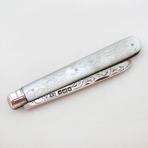 May include: A silver-toned antique penknife with a mother-of-pearl handle. The handle is engraved with floral patterns. The blade is open, revealing intricate scrollwork. The penknife has a silver cap on one end and hallmarks.