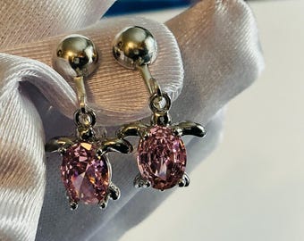 May include: A pair of silver-toned drop earrings featuring a turtle-shaped design. Each earring has a polished metal head and a faceted oval-shaped pink gemstone forming the body of the turtle, suspended from a small round silver-toned stud.