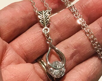 May include: A silver-toned necklace featuring a pendant with an arrow-shaped top and a teardrop-shaped loop at the bottom. A clear, round, faceted stone is set within the teardrop loop. The pendant hangs from a delicate silver-toned chain link necklace.
