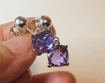 May include: A pair of silver-toned earrings featuring cushion-cut, faceted purple stones. Each earring is secured with a polished silver-toned ball-style screw-back mechanism, showcasing a clear, vibrant violet hue and a reflective, multi-faceted surface.