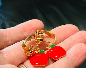 May include: Gold-toned clip-on earrings featuring a bright red apple design with green leaves. The apples have a glossy finish, and the clip mechanism is visible. These earrings are a fun, playful accessory.