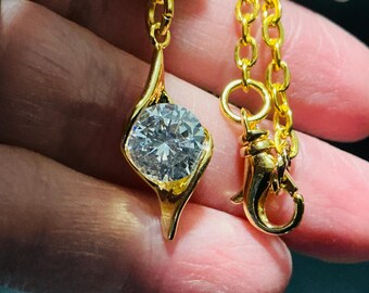 May include: A gold-toned necklace featuring a single round, clear, faceted stone set within a swirling, teardrop-shaped metal frame. The pendant is attached to a delicate gold-toned chain that includes a small lobster claw clasp for closure.