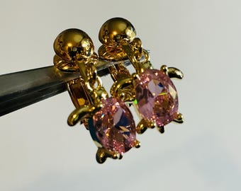 May include: A pair of gold-colored earrings featuring a turtle design. Each earring has a pink, oval-shaped gemstone set within the turtle's body. The earrings are held by a metal tool against a white background.