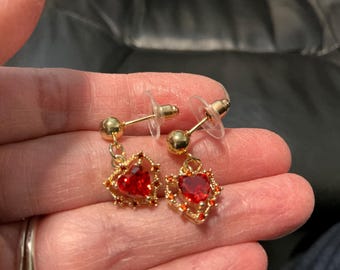 May include: A pair of gold-colored earrings with red heart-shaped pendants. The earrings have a post-and-stud design with a small gold ball at the top. The heart pendants are surrounded by a decorative gold frame.