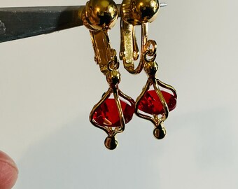 May include: Clip-on earrings with a gold-tone metal design. Each earring features a cage-like structure holding a faceted, deep red gemstone. The earrings have a classic, elegant style, suitable for formal occasions.