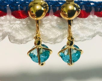 May include: A pair of gold-tone clip-on earrings featuring round, faceted, bright blue stones held in a cage-like setting. The earrings are suspended from a gold-tone circular clip mechanism and are displayed against a white, red, and blue trim.
