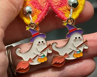 May include: A pair of clip-on earrings featuring white ghost charms wearing purple witch hats with red bands. Each ghost holds a brown broomstick and a small orange pumpkin bucket filled with colorful candy. The charms are attached to yellow and pink fabric.