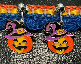 May include: A pair of silver-toned clip-on earrings featuring a dangling enamel charm of a jack-o'-lantern pumpkin wearing a purple and black witch hat with a yellow buckle. The pumpkin is orange and yellow with a black carved face design.