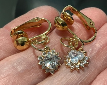 May include: A pair of gold-toned clip-on earrings. Each earring features a central, clear, faceted stone surrounded by smaller, clear stones, creating a floral or starburst design. The clip mechanism is visible, and the earrings are held in a hand.