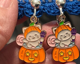 May include: A pair of silver-tone clip-on earrings featuring a white cat character wearing an orange pumpkin hat and sitting inside a carved orange pumpkin base with yellow star cutouts and a small purple lollipop held in one paw.