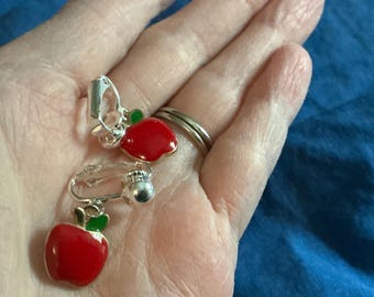 May include: A pair of clip-on earrings featuring a red apple design with green leaves. The earrings have a silver-colored metal clip. The earrings are shown against a blue background.