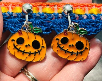 May include: A pair of silver-toned clip-on earrings featuring orange pumpkin charms with black enamel eyes and stitched-style mouths, accented with small green leaves at the top. The pumpkin charms are suspended from polished silver-toned metal clips.