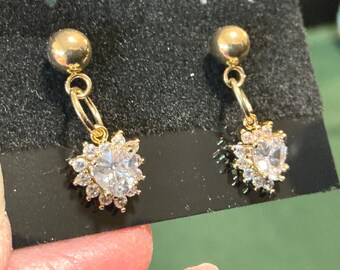 May include: A pair of gold-colored earrings. Each earring features a small, round stud connected to a dangling charm. The charm is a heart-shaped, clear crystal surrounded by smaller, clear crystals, creating a floral design.