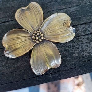 May include: A detailed, antique-style brass flower with four petals and a textured center. The flower is a warm gold color and appears to be made of metal. The petals have a slight curve and are detailed with lines. The flower is a decorative item.