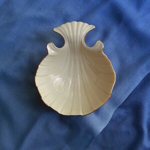 May include: Cream-colored, shell-shaped dish with a gold trim. The dish has a scalloped design and a flared top. The dish is likely made of porcelain or ceramic and is a decorative item.