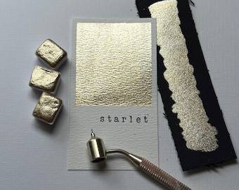 May include: A collection of gold watercolor paint samples and tools. The image features three small, square paint cubes, a swatch on black paper, and a sample card labeled "starlet". A metal pen-like tool is also present.