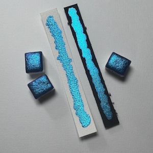 May include: Two swatches of teal blue metallic watercolor paint on white paper, next to two teal blue watercolor pans.