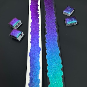 May include: Two strips of iridescent paint, one on white paper and one on black paper, with two small pans of iridescent paint labeled "galactic" and "phoebe".