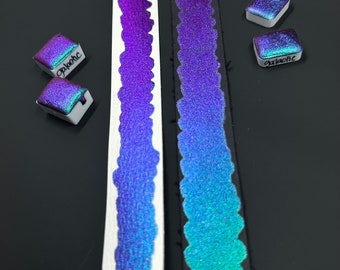 Könnte beinhalten: Zwei Streifen irisierender Farbe, einer auf weißem Papier und einer auf schwarzem Papier, mit zwei kleinen Dosen irisierender Farbe mit den Bezeichnungen "galactic" und "phoebe".