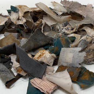 Könnte beinhalten: Eine Ansammlung verschiedener Stoffreste in unterschiedlichen Formen und Größen. Die Stücke weisen eine Mischung aus Farben wie Petrol, Braun, Grau, Creme und Weiß auf, wobei einige abgenutzte Texturen, ausgefranste Kanten und Muster zeigen.