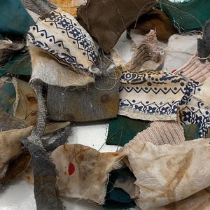 May include: A collection of assorted fabric scraps in various shapes and sizes. The pile includes pieces of dark green, grey, and off-white cloth, some featuring blue and white patterned prints, with visible frayed edges and distressed textures.