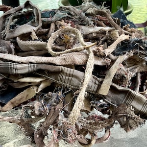 May include: A pile of assorted fabric scraps in various shades of brown, gray, and blue. The scraps include strips of plaid, denim, and other textured materials, with frayed edges and a rustic appearance. The fabrics are bunched together.