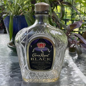 Puede incluir: Una botella de vidrio transparente Crown Royal Black con un patrón de diamantes. La etiqueta presenta un diseño de corona y el texto "Crown Royal Black, Blended Canadian Whisky". La botella tiene un tapón de corcho.