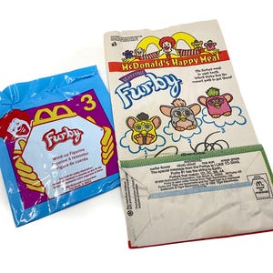 May include: A blue plastic McDonald's Happy Meal bag featuring Ronald McDonald and the text Furby Wind-up Figurine, alongside a white paper bag with a Furby maze game, the McDonald's logo, and printed text including 1998 McDonald's Corporation.