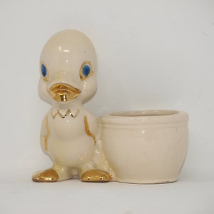 May include: A white ceramic planter shaped like a duck with gold accents and blue eyes. The planter has a separate pot for planting.