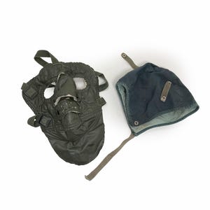May include: An olive green quilted face mask with eye openings and adjustable straps alongside a dark blue cap featuring ear flaps, a chin strap, and a small rectangular patch on the side. Both items are constructed from fabric with visible stitching.