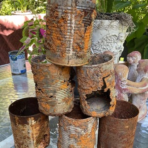 May include: A stack of six cylindrical metal cans arranged in a pyramid formation. Each can features a heavily rusted, textured surface with patches of orange and brown oxidation, showing significant wear, jagged edges, and holes in the metal.