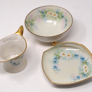 Puede incluir: Un juego de tres platos de porcelana blanca con un diseño floral. Los platos presentan un delicado patrón de margaritas blancas y nomeolvides azules con hojas verdes. Los platos tienen un borde dorado.