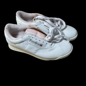 Op de afbeelding: Paar witte Reebok sneakers met veters, een laag profiel en een gewatteerd interieur. De schoenen hebben een klassiek design met het Reebok-logo aan de zijkant en een crèmekleurige zool. De sneakers staan tegen een zwarte achtergrond.