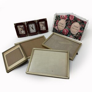 May include: A collection of picture frames including a triple-hinged dark red frame with a central clock, a rectangular fabric-covered frame with floral patterns and two oval openings, and three rectangular gold-toned metal frames.