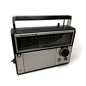 Puede incluir: Una radio portátil Realistic Patrolman CB-60 en color negro y plata con asa superior, rejilla de altavoz frontal, un dial de sintonización grande y un panel de visualización de frecuencias con bandas AM, FM, CB y VHF en texto blanco y azul.