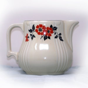 May include: A white ceramic pitcher with a floral design. The pitcher has a handle and a spout. The flowers are red with black centers and are on a vine with green leaves.