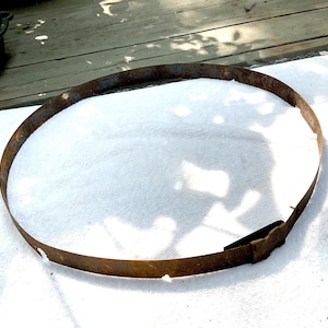 May include: A circular, rusty metal band, possibly from a barrel, lies on a white surface. The aged hoop displays a dark brown hue and signs of wear and tear, indicative of its use and exposure to the elements.