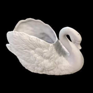 Puede incluir: Una maceta o cuenco decorativo de cerámica blanca con forma de cisne, que presenta texturas de plumas moldeadas detalladas en todo el cuerpo. El cisne tiene un cuello curvado y un centro hueco y abierto, con un acabado mate y liso.